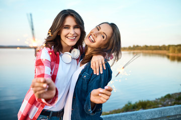 Two beautiful young women smiling and staring at camera holding sparklers with view of lake in background