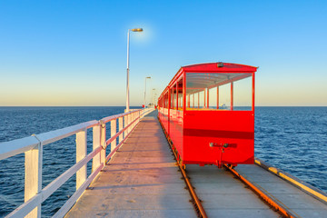 Wall Mural - View from behind of vintage red train illuminated by sunset light, going on Jetty Busselton tracks in Busselton town, WA. Busselton Jetty is the longest wooden pier in the world and iconic place.