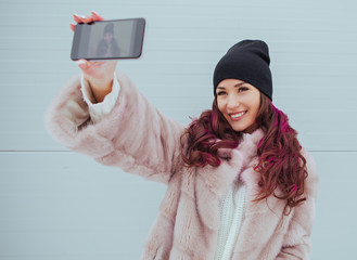 Wall Mural - Fashion portrait of pretty smiling hipster woman in mink coat and smartphone against the colorful grey wall. Making selfie. Casual clothes. Colored hairs