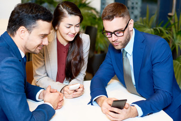 Portrait of three young business people using smartphones sitting at table in cafe, looking at screen and smiling