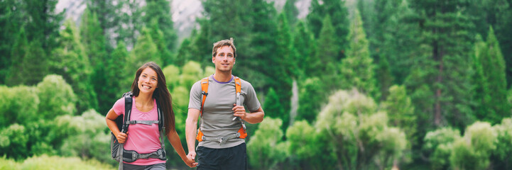 Wall Mural - Happy hikers couple walking in green forest hiking in nature landscape background. Multiracial group of young people living an active lifestyle panorama banner. Asian girl, Caucasian man.