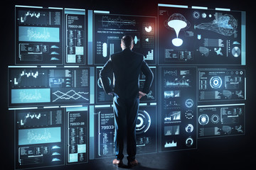 Thoughtful programmer. Calm concentrated experienced programmer standing in front of a giant futuristic device with his hands on the hips and thoughtfully looking at the new information