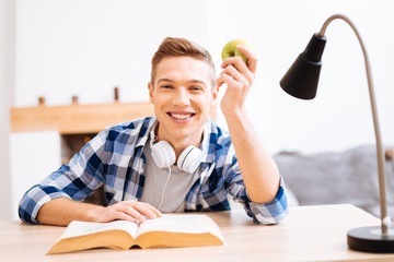 Wall Mural - I love fruit. Good-looking delighted fair-haired boy smiling and sitting at the table with a book and holding an apple