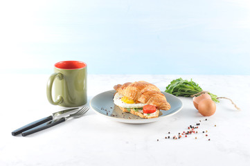 Croissant with arugula, fried egg, slice of tomato. In the frame is a cup of coffee, a bunch of arugula and cutlery. Light background.