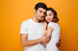 Photo of masculine man in casual white t-shirt hugging his wife while standing on camera, isolated over yellow background