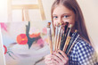 © Viacheslav Yakobchuk - SEt of a real artist. Positive girl sitting at her easel and holding many already used painting brushes while smiling and posing for the camera.