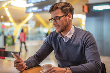 young man at the airport using his mobile phone and drinking coffee waiting for his flight close-up portrait