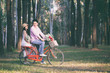 A lover couple ride a bike in a pine forest, woman has a bunch of flowers in her hands.