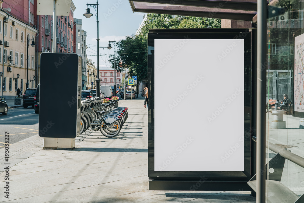 Vertical blank white billboard at bus stop on city street. In the background buildings and road. Mock up. Poster on street next to roadway. Sunny summer day. - obrazy, fototapety, plakaty 