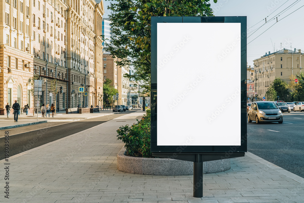 Vertical blank glowing billboard on the city street. In the background buildings and road with cars. Mock up. The poster on the street next to the roadway. - obrazy, fototapety, plakaty 