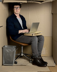 Too closely concept. Full length portrait of confused businessman is sitting in cramped carton office while holding modern laptop on his knees. He is looking at camera disappointedly