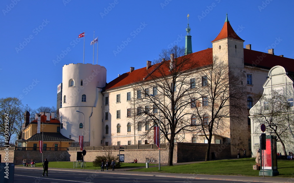 Obraz na płótnie Zamek w Rydze, stolicy Łotwy, mały park przed budowlą, ludzie spacerują, siedzą na ławkach i na trawie, słoneczny dzień, błękitne bezchmurne niebo w salonie