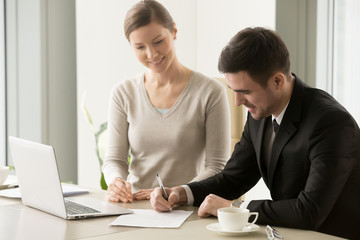 Wall Mural - Happy male company head signing contract sitting at desk in office with smiling female business partner. Businessman putting signature on agreement. Young businesswoman satisfied with closing the deal