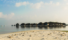 In Foreground: A Beach With Sandy Hills Made By Crabs, In Background: Beautiful Wooden Villas, Standing On Stilts In Water Of Indian Ocean, Sunrise, Maldives. Travel And Tourism Concept