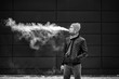Vape man. Portrait of a handsome young white guy in casual clothes vaping an electronic and letting out puffs of steam cigarette opposite the futuristic modern building. Close up. Black and white.