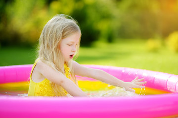 Wall Mural - Adorable little girl playing in inflatable baby pool. Happy kid splashing in colorful garden play center on hot summer day.