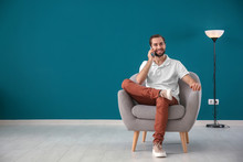 Handsome Man Talking On Mobile Phone While Sitting In Comfortable Armchair Against Color Wall