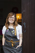 © Mint Images - Woman with long brown hair wearing apron and glasses standing in an open doorway, smiling.