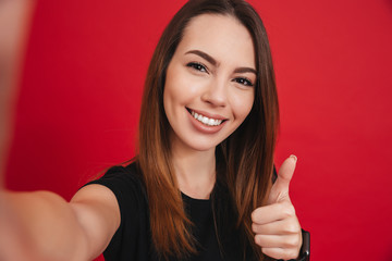 Sticker - Young happy woman 20s with long brown hair smiling and taking selfie with gesturing thumb up, isolated over red background