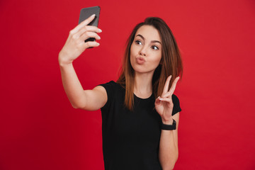 Sticker - Lovely woman in black t-shirt having fun and taking selfie with gesturing peace sign on her smartphone, isolated over red background