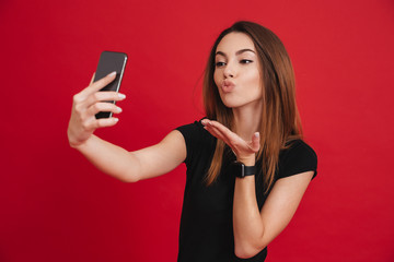 Sticker - Photo of modern woman in black t-shirt partying and making air kiss on mobile phone isolated over red background