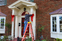 Woman On Ladder Putting Christmas Decorations Up On Pillars Of Entrance Of Two Story Brick House With Bay Windows
