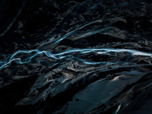 Aerial Of An Abstract Blue River In Iceland In A Contrast Black Sand Beach Riverbed