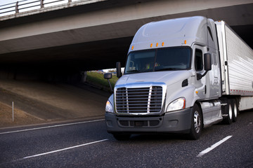 Amazing big rig grey semi truck with semi trailer driving under bridge across the highway