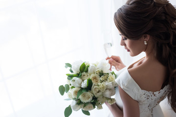 attractive bride in traditional dress with wedding bouquet and glass of champagne standing at window