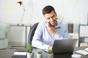 Wall Mural - Young man talking on phone while working with laptop in office