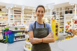Fototapeta  - Happy natural young saleswoman in modern supermarket. Cross arms. Looking camera
