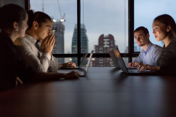 Business people team working late night in low light from laptop screen with cityscape blurred background.Overtime hours concept.