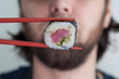 Hand using chopsticks pick. Maki-Sushi, tuna-maki rolls in front of face of young man. Fresh made Sushi set with tuna, cheese and cucumber. Traditional Japanese cuisine.