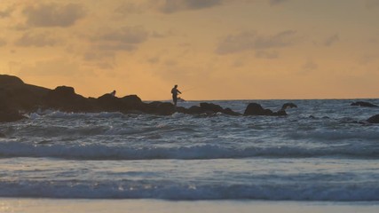 Wall Mural - silhouette of a fisherman on rocks against sunset sea in slow motion