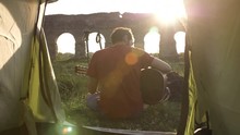 Young Man Backpacker Playing Guitar Outside Camping Tent Sitting On Grass In Front Of Roman Aqueduct Arches In Parco Degli Acquedotti Park Ruins In Rome At Sunset Sticks In Ground Camera Shot Inside