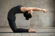 Young woman practicing yoga, doing Preparation for kapotasana, variation of Ustrasana exercise, Camel pose, working out, wearing sportswear, black pants and top, indoor full length, gray wall, studio