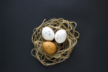 Nest with fresh chicken egg with drawn faces on black paper background. Concept for Easter with copy space. Creative layout made of white and brown eggs. Top view photo.