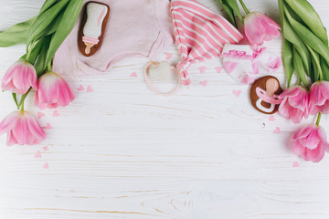 A composition for newborns on a wooden white background with clothes, pink tulips, hearts and a cookies, copy space and flat lay. It's a girl.