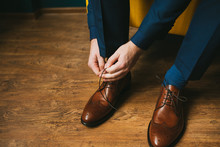 A Man Or A Groom In A Blue Suit Ties Up Shoelaces On Brown Leather Shoes Brogues On A Wooden Parquet Background