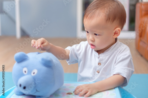 Cute Little Asian 18 Months 1 Year Old Toddler Baby Boy Child Cute Little Asian 18 Months 1 Year Old Toddler Baby Boy Child Putting Thai Coin Into Blue Piggy Bank Money Savings For Children Financial