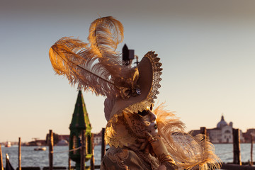 VENICE, ITALY - FEBRUARY 10 2018: Close up of golden dressed carnival mask adorned with fantasy motifs. The masks are photographed with the background of the Giudecca cana