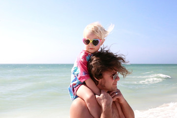 Little Toddler Girl Sitting on Father's Shoulders on Beach by the Ocean