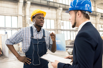 Wall Mural - Bearded African American factory worker wearing hardhat and overall discussing results of accomplished work with his superior while standing at production department