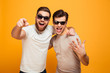 © Drobot Dean - Two happy men friends in casual t-shirts and sunglasses having fun together and pointing fingers on camera meaning hey you, isolated over yellow background