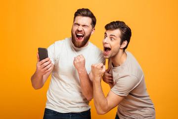 Wall Mural - Image of two bachelors screaming and rejoicing win of football team while watching game on mobile phone, isolated over yellow background