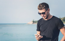 Handsome Man In Sunglasses Holding Mobile Phone In His Hand And Texting Message, On Tropical Background.