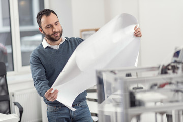 Auspicious beginning. Charming bristled young engineer standing in the office and examining the blueprint in his hands, being satisfied with the result