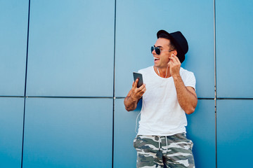Wall Mural - Profile portrait of a cheerful young man in sunglasses, listening to music in headset on mobile phone. Dressed in fashionable clothes. Outdoors. Copy space