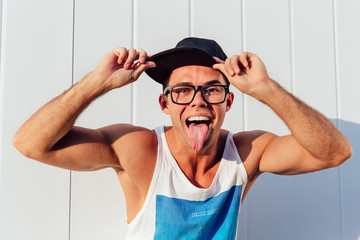 Wall Mural - Cheerful muscular guy in eyeglasses and cap showing a tongue at camera, while having fun outdoors. Dressed in stylish singlet.