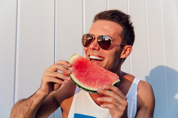 Wall Mural - Handsome young man in sunglasses eating a watermelon, enjoying the summer days, outdoors. Dressed in sleeveless t-shirt.